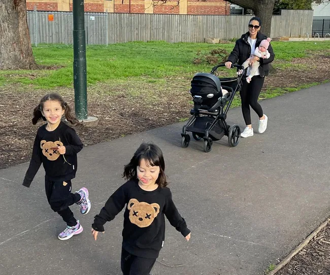 Woman pushes a stroller with a baby, while two children in matching bear sweaters run ahead on a path.
