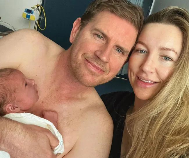 Couple smiling with a newborn baby in a hospital room, man is shirtless holding the baby.