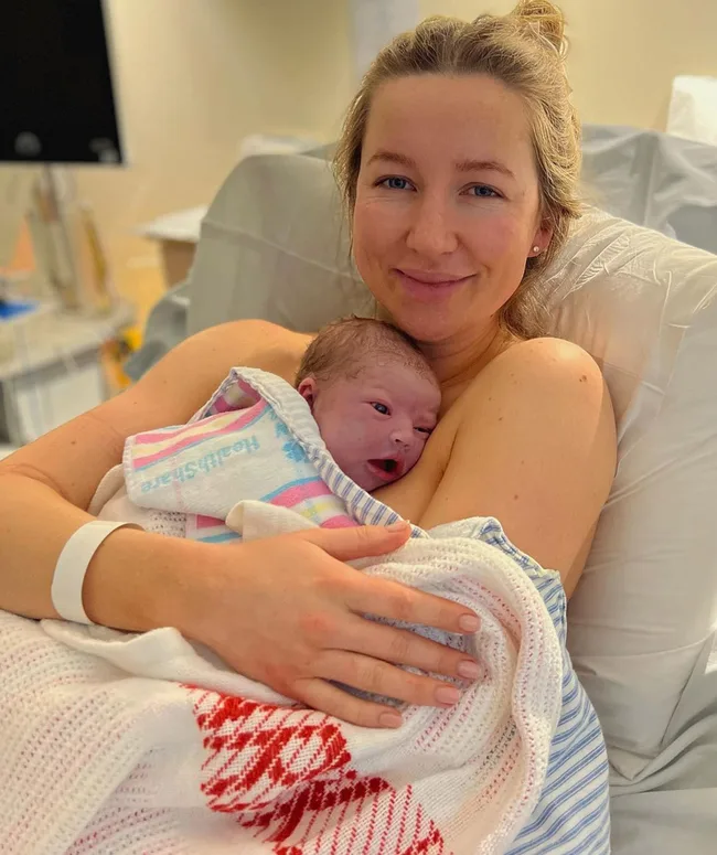 A woman smiling and holding a newborn wrapped in a hospital blanket, resting in a hospital bed.