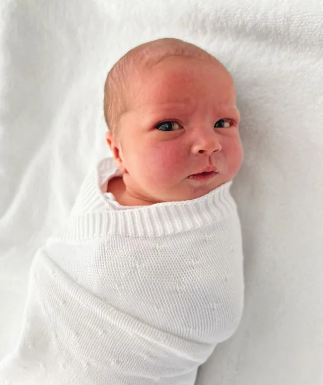 A newborn baby wrapped in a white knitted blanket, lying on a white surface, with a curious expression.
