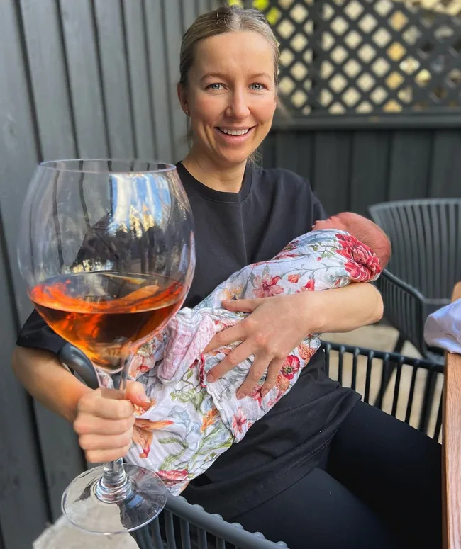 A woman smiles while holding a swaddled baby and a large glass of orange drink outdoors.