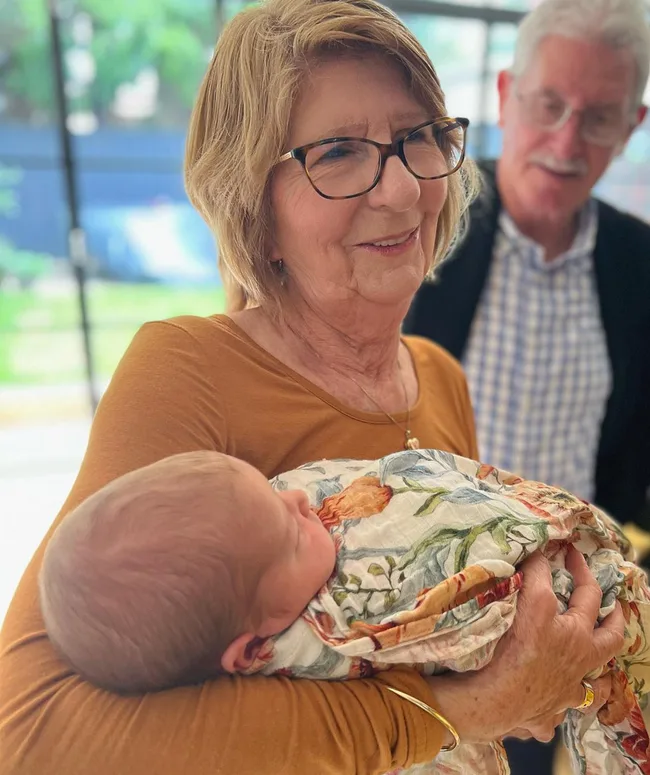 Grandmother holding a swaddled newborn baby with a floral blanket, while a man stands smiling in the background.