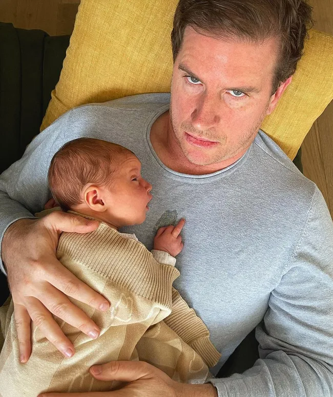 A man lying on a couch holding a sleeping baby wrapped in a beige blanket on his chest.