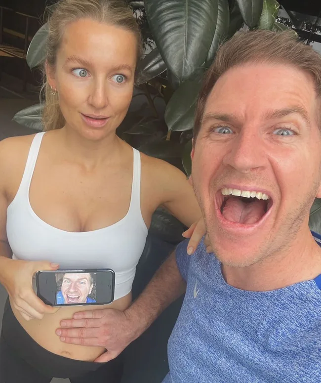 A man and woman pose playfully; the woman holds a phone with the man's face on her belly, both smiling widely.