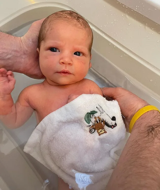 A newborn baby in a bath is gently supported by an adult's hands, covered partially with a towel featuring a monkey design.