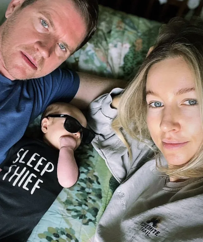 Man and woman lying on bed with baby wearing sunglasses and "SLEEP THIEF" shirt.