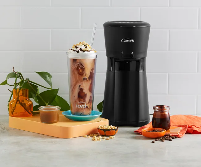 Sunbeam coffee maker with iced coffee, whipped cream, and ingredients on a kitchen counter.