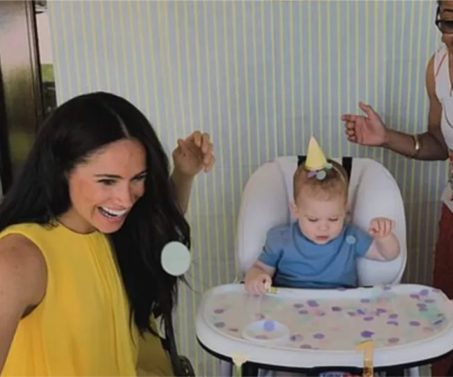Woman in yellow smiling at a seated baby wearing a party hat, with balloons and confetti around.