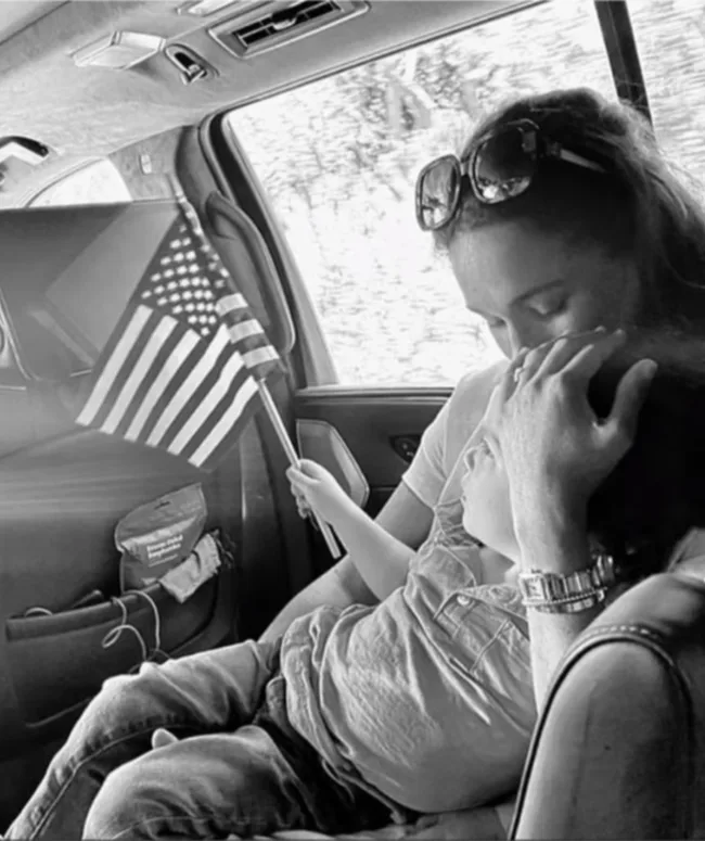 A woman in a car gently holds a child resting on her lap, who is holding an American flag.
