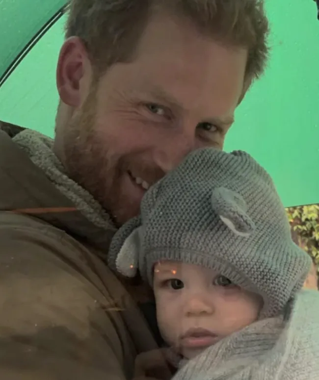 A man smiling while holding a baby wearing a knitted hat with bear ears under a green umbrella.