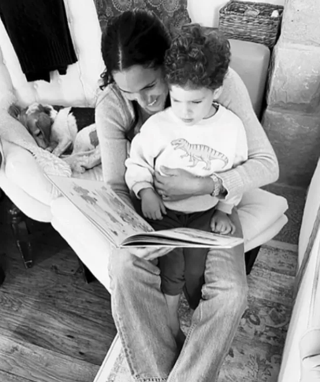 A woman sits with a child on her lap, reading a book together in a cozy room. A dog sleeps on a chair nearby.