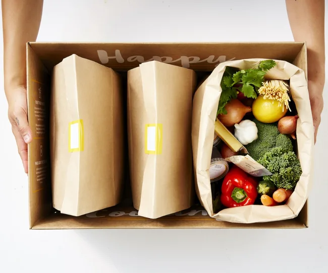 A box with brown bags and fresh vegetables including broccoli, red pepper, lemon, onion, and herbs, held by two hands.