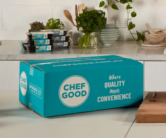 Blue Chefgood box on a kitchen counter with stacked meal trays and herbs in the background.