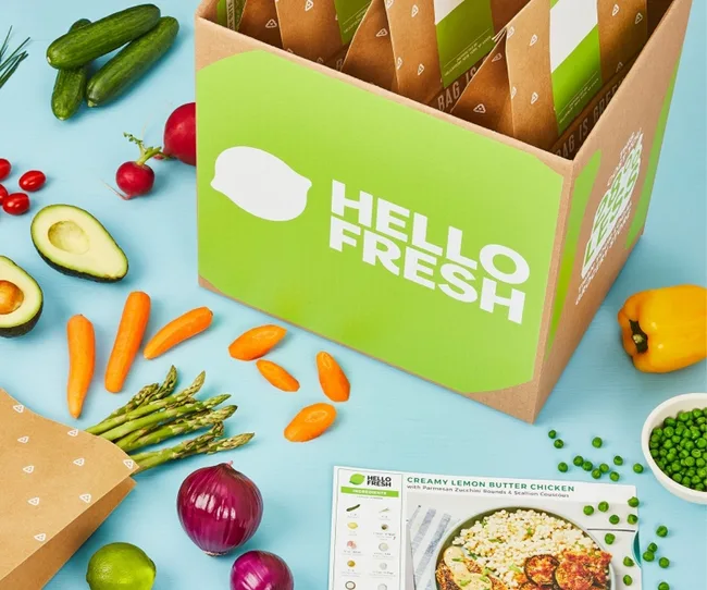 Box with HelloFresh logo surrounded by fresh vegetables and a recipe card on a blue background.