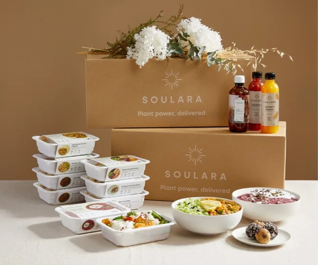 Alt text: "Soups, bottles, and bowls of plant-based meals with Soulara packaging on a table, flowers on boxes in background."