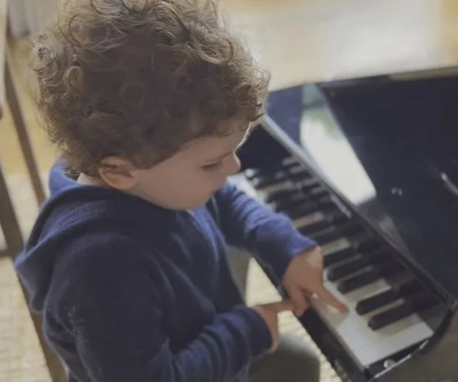 Young child with curly hair playing a small piano, wearing a navy blue sweater, focused on the keys.