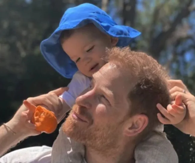 Father carrying a young child in a blue hat on his shoulders, holding a peeled orange, outdoors.