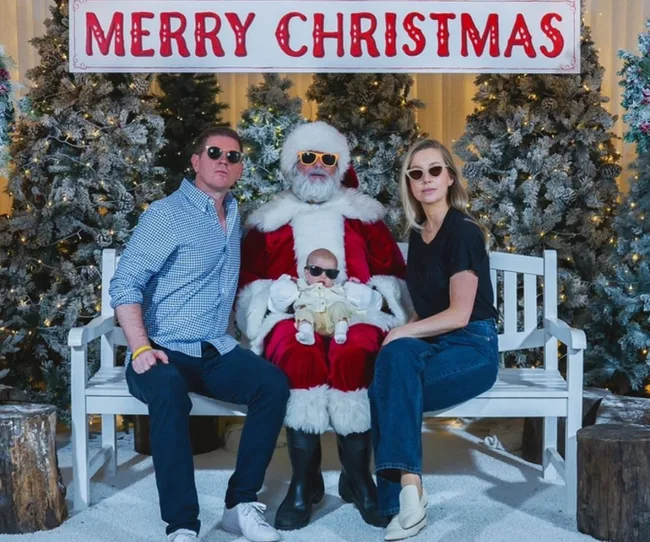 A family with sunglasses poses with Santa Claus under a "Merry Christmas" sign, surrounded by snowy trees.