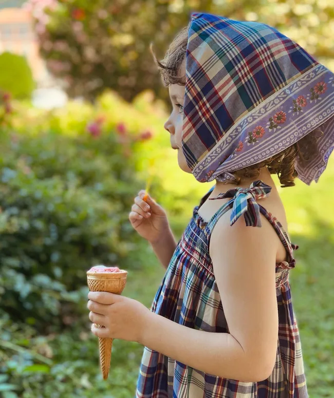 A young girl in a plaid dress and headscarf holds an ice cream cone with a garden in the background.