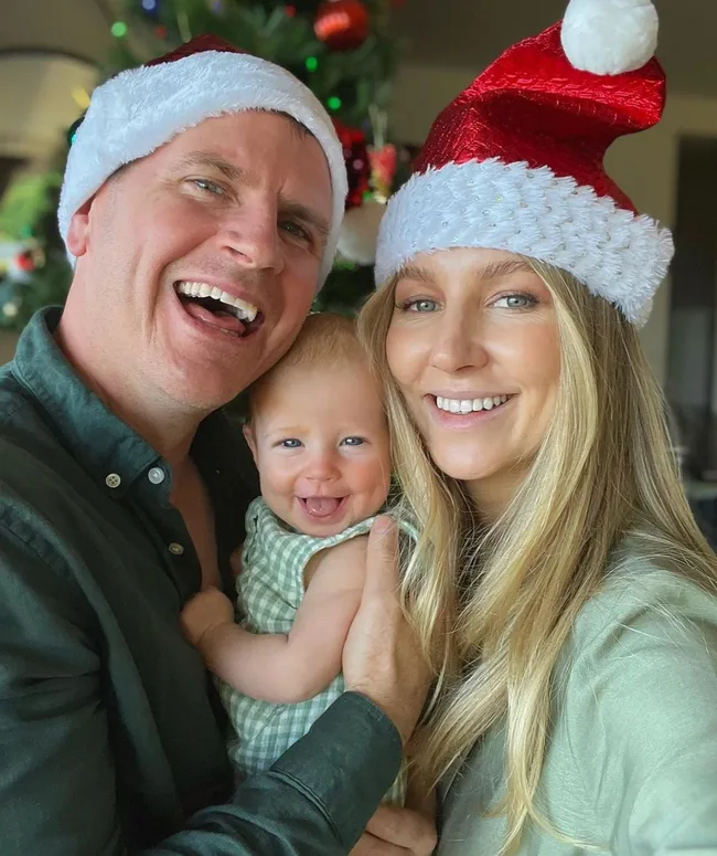 Man and woman wearing Santa hats smiling with a baby in front of a Christmas tree.