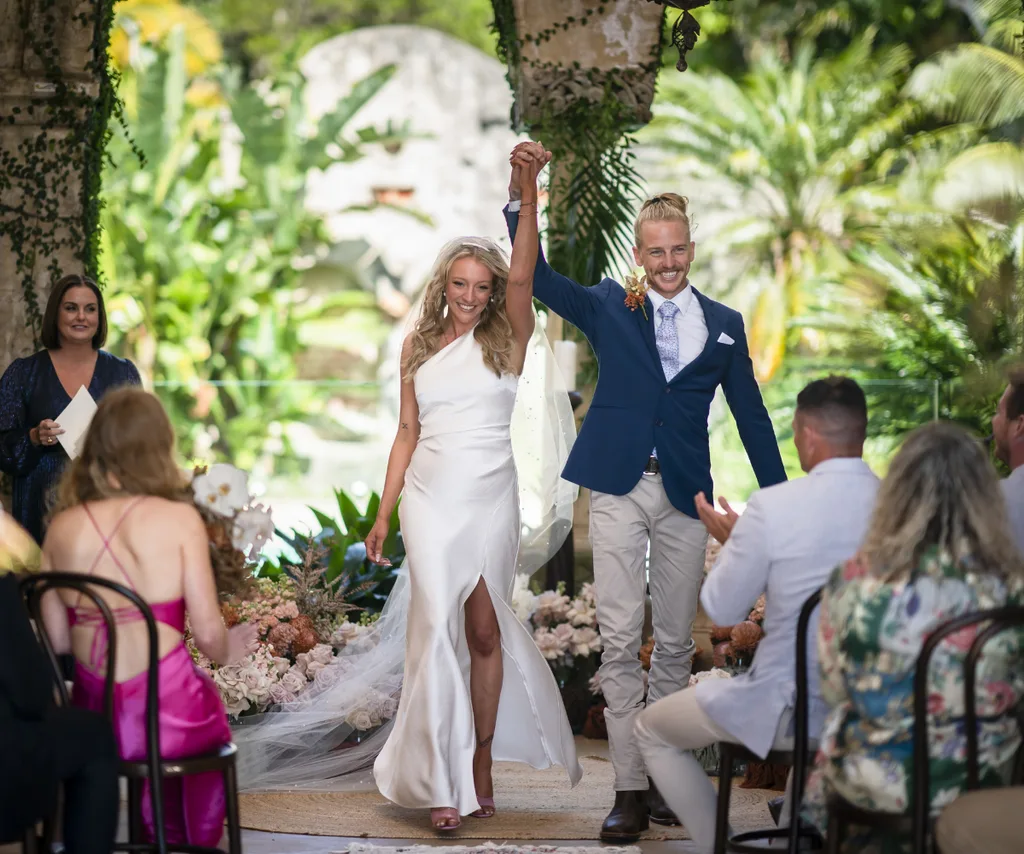Couple celebrates at garden wedding, bride in white dress, groom in blue suit, surrounded by lush greenery and guests.