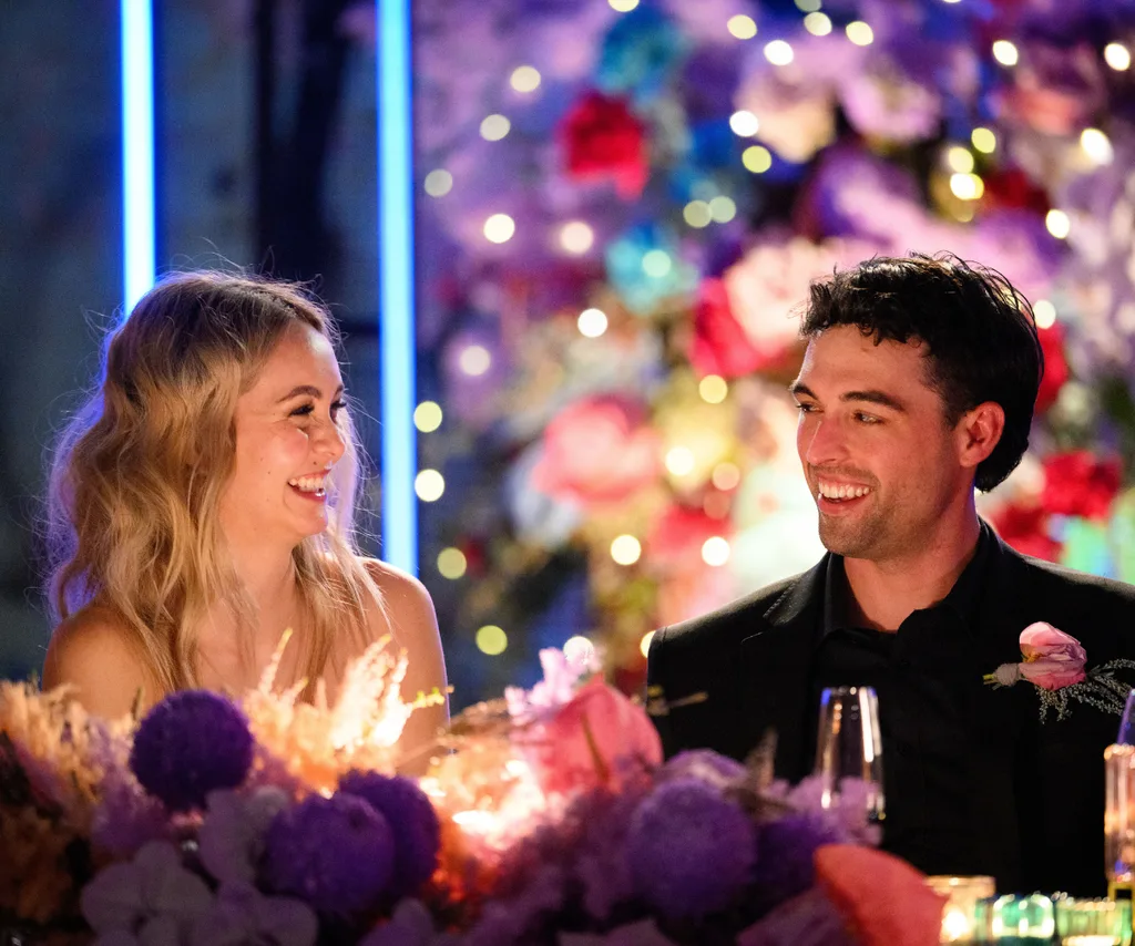 Two people smiling at each other at a colorful, flower-decorated event.