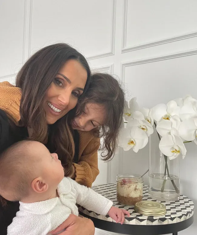 A woman smiles with two young children, one baby in her arms, near a table with white flowers and a jar.