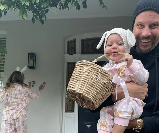 Man holding a smiling baby wearing bunny ears, with another person in pajamas in the background near house number 17.