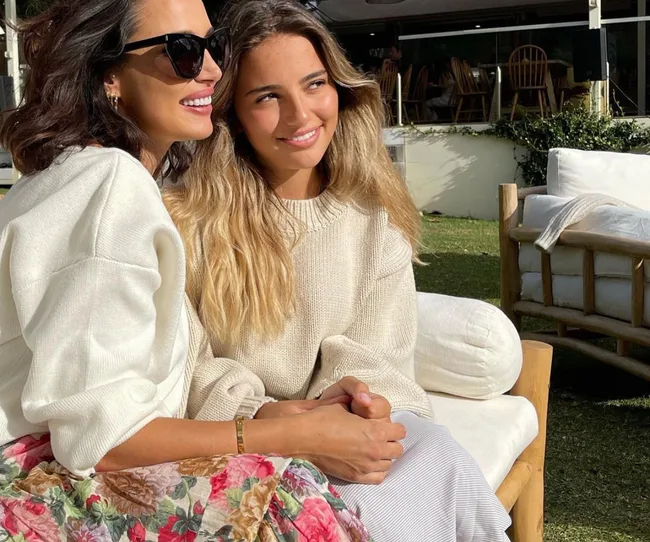 Two women sitting together outdoors, smiling, wearing light sweaters. One has sunglasses, and there's a floral skirt.