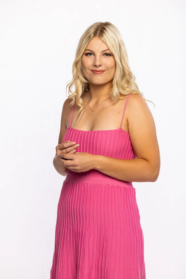 Blonde woman in a sleeveless pink dress with a slight smile, hands clasped, against a plain white background.