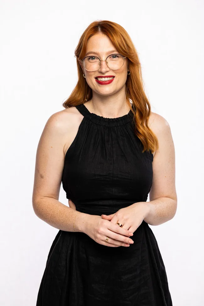 Red-haired woman in glasses and a black dress smiles against a plain white background.