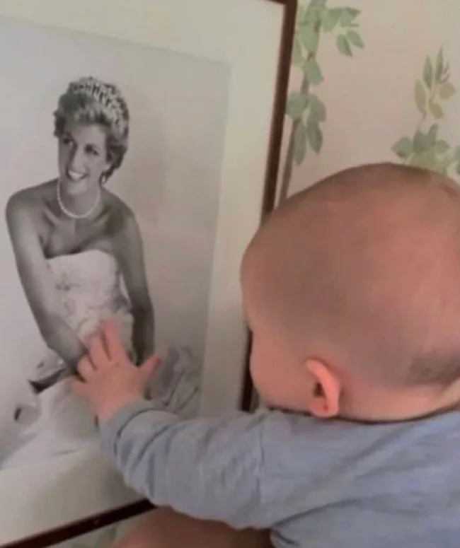 ```html
A baby reaching out to touch a framed photograph of a smiling woman in a tiara and gown.
```