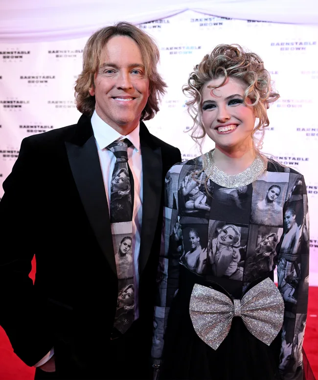 Father and daughter smiling on the red carpet, daughter wearing dress with vintage portraits and large bow.