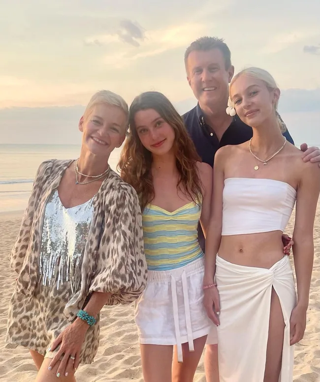 A family of four smiling at the beach during sunset, dressed in summer clothing.