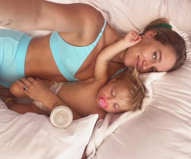 Mother in blue top cuddling with sleeping baby with a pacifier, lying on a bed.