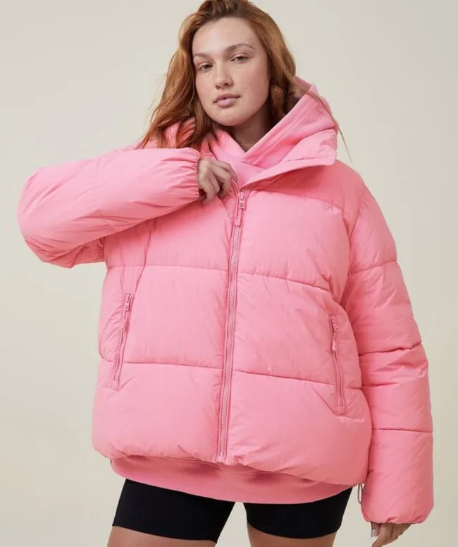 A person wearing a pink puffer jacket and black shorts, posing with one hand near the collar.