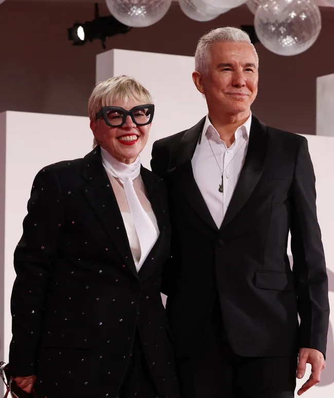 Two people pose together; one wearing glasses and a tuxedo with a white scarf, the other in a classic black suit and white shirt.