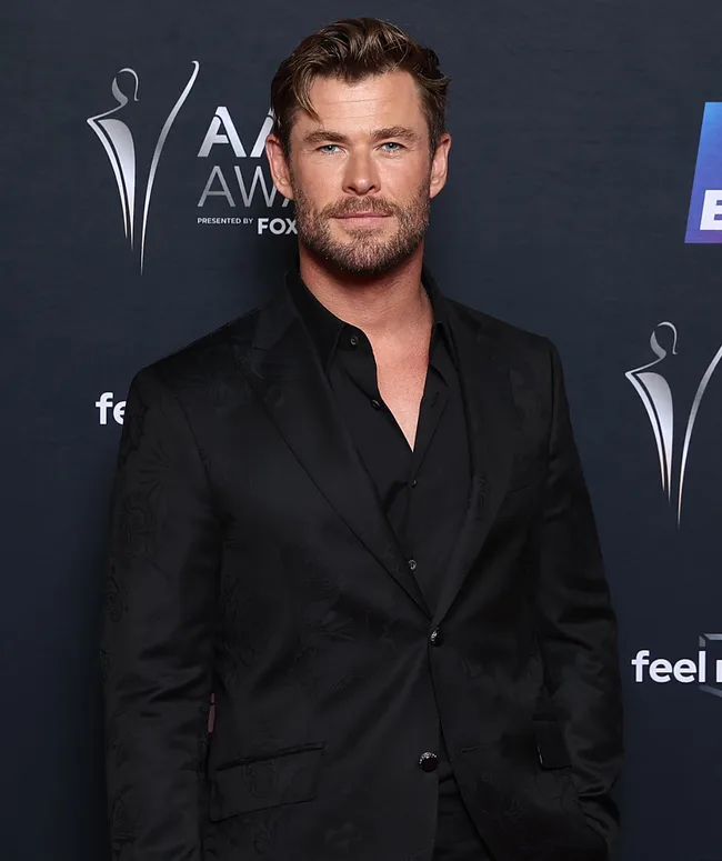 Man in a dark floral suit posing at an awards event with a dark backdrop featuring stylized statues.