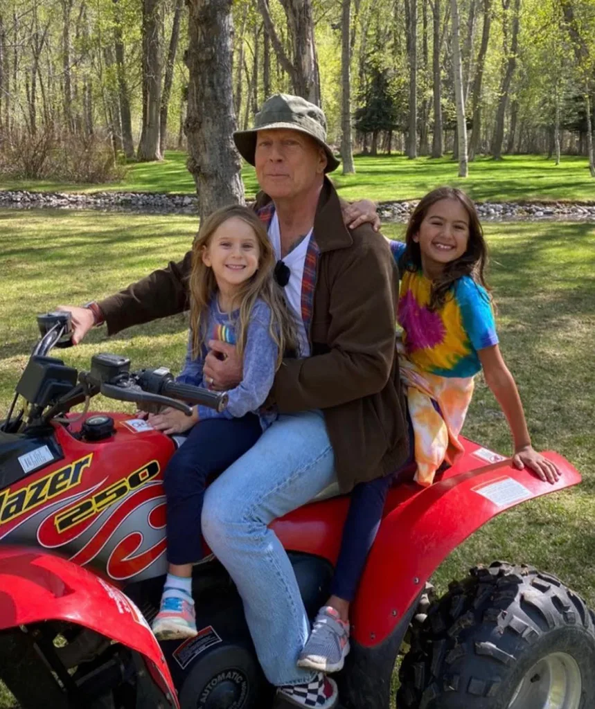 Man wearing a hat on a red ATV with two smiling children, surrounded by green trees and grass.