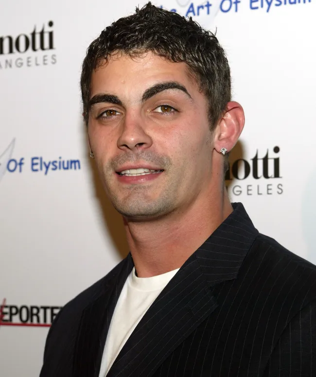 Man in a suit with stud earrings, smiling at an event backdrop featuring logos.