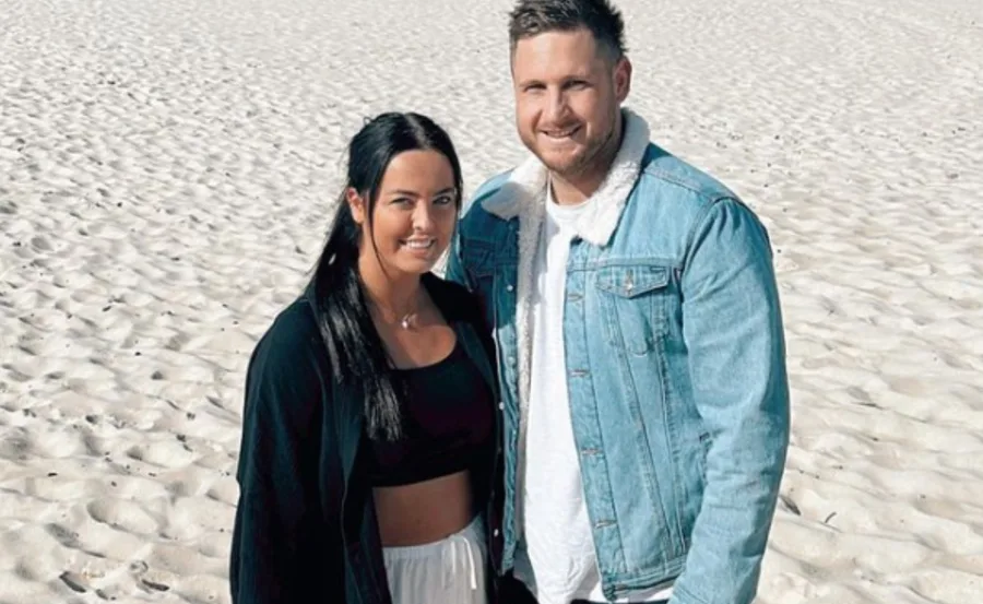 A couple standing on a sandy beach, smiling, both wearing casual outfits; the man in a denim jacket.