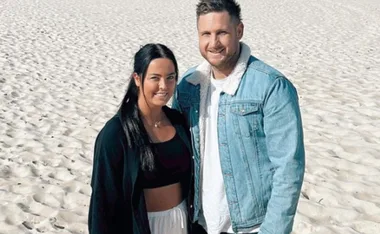 A couple standing on a sandy beach, smiling, both wearing casual outfits; the man in a denim jacket.