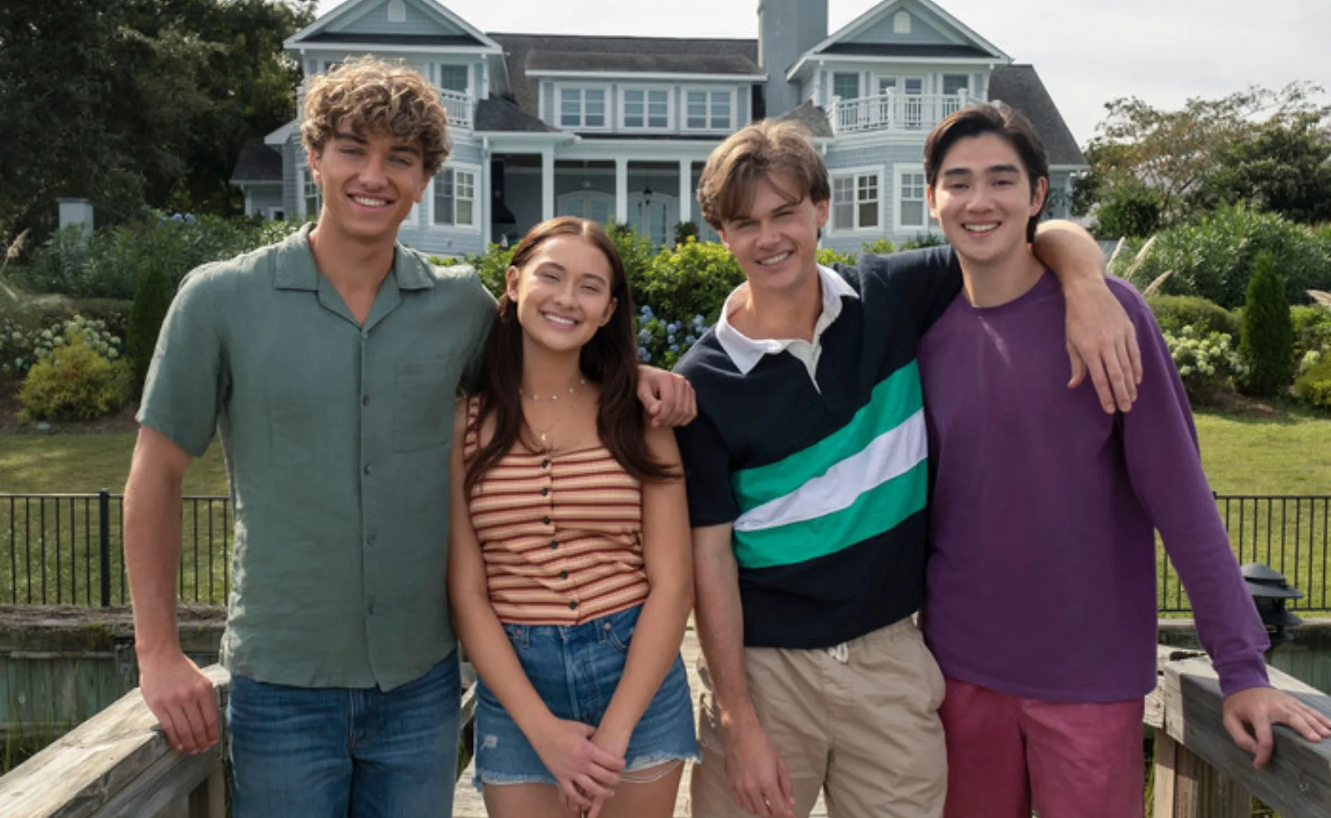 Group of four young adults smiling outside, standing arm in arm on a wooden bridge with a large house in the background.