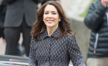 A smiling woman with brown hair in a patterned coat walks outside; people and a building in the background.