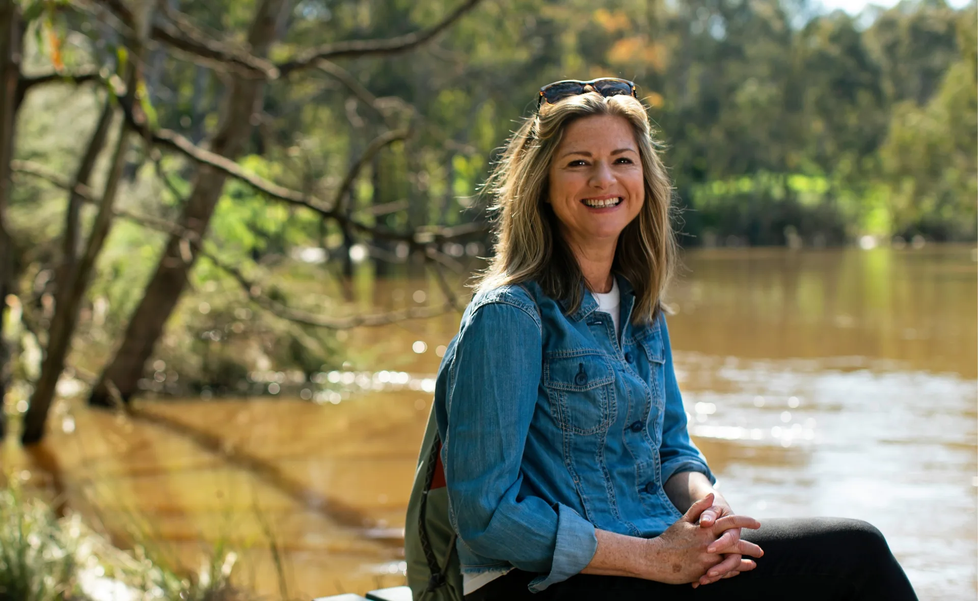 Woman in denim shirt smiling by a calm, forested riverbank, wearing sunglasses on her head.