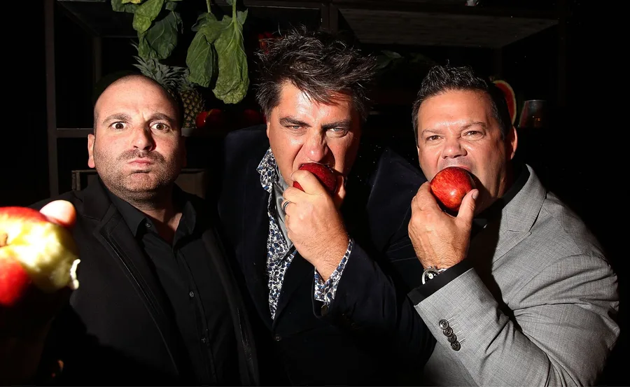 Three judges holding and biting apples, looking directly at the camera with dramatic expressions.