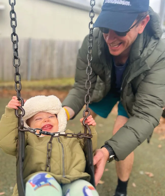 Smiling baby in winter clothes on a swing, gently pushed by a man in casual attire at a park.