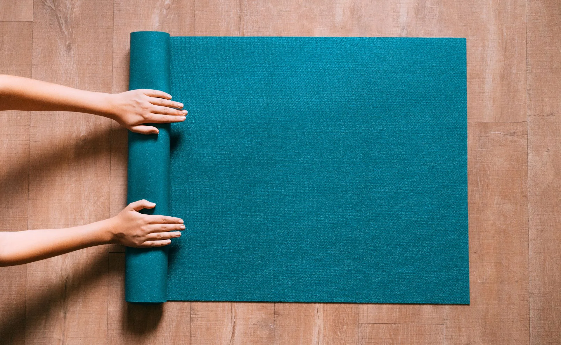 Person's hands rolling a teal Pilates mat on a wooden floor.
