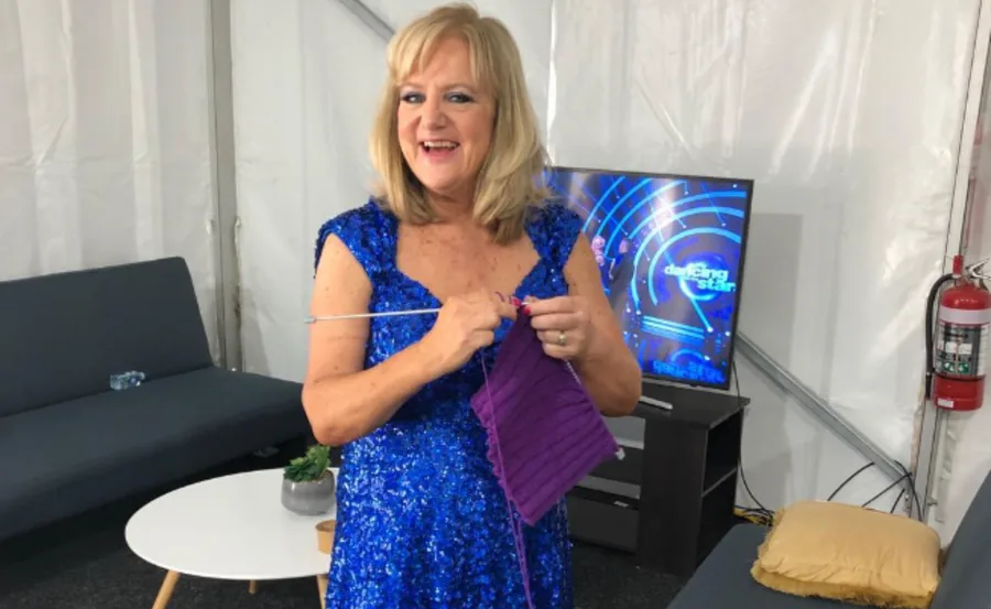 Woman in a sparkly blue dress knitting purple fabric, standing near a sofa with "Dancing with the Stars" on TV.