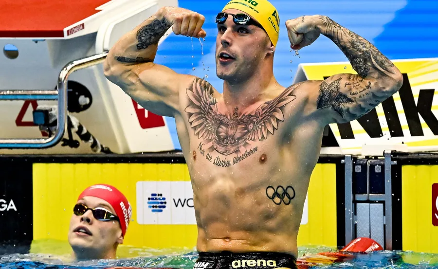 Swimmer flexes muscles in pool with tattoos and Olympic rings, wearing a yellow cap; another swimmer looks on.